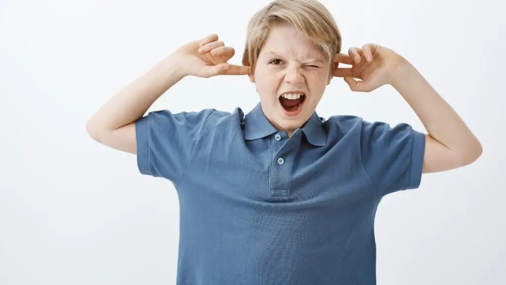 How to deal with overstimulation autism: a boy in a blue shirt covers his ears and has one eye closed.
